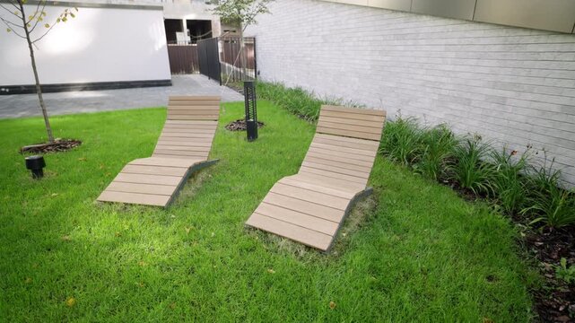 Wooden chaise lounges rest on green lawn near rustic brick wall. Sunbathing area offers peaceful relaxation spot in residential garden