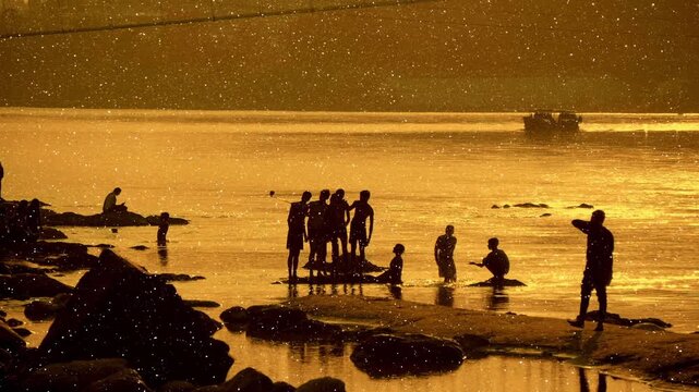 Indian boys take selfies on a smartphone and grown men swim and play with joy on the rocks in the waters of the Great Ganges (Ganga), bathed in the golden rays of the setting sun. A boat is sailing on