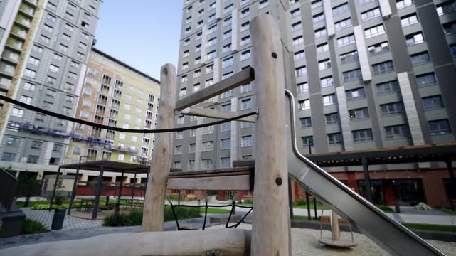 Wooden playground structure includes metal slide in sandy area. Apartment buildings provide contrasting background to natural kids zone