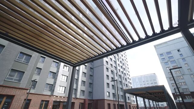 Striped wooden roof filters sunlight through narrow parallel slats. Architectural shadows fall across pavement in residential district
