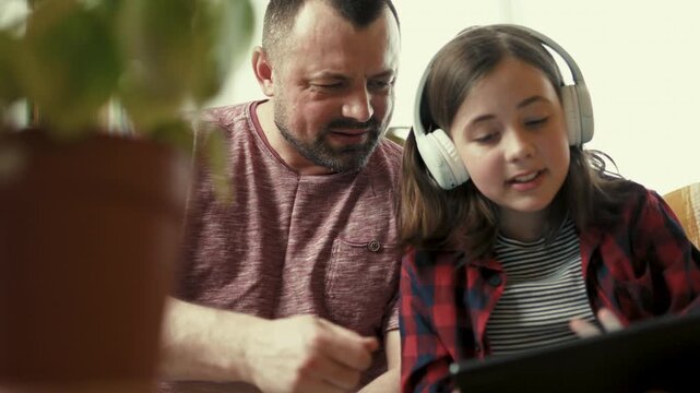 Preteen girl in headphones showing interesting content on digital tablet to her father at home-4k,10 bit footage.