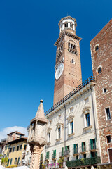 Fototapeta premium Historic clock Lamberti Tower in Verona, Italy, featuring intricate architecture and a clear blue sky, surrounded by traditional buildings and a decorative column