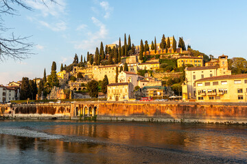 Naklejka premium Scenic view of historic Castel San Pietro and lush greenery on a hillside in Verona, Italy, with the Adige River reflecting the warm sunset light