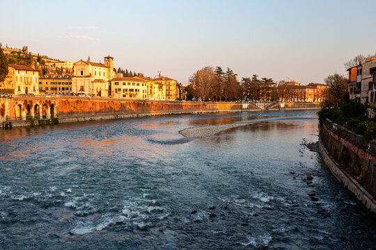 Scenic view of the Adige River flowing through Verona, Italy, with historic buildings and trees lining the riverbank during sunset