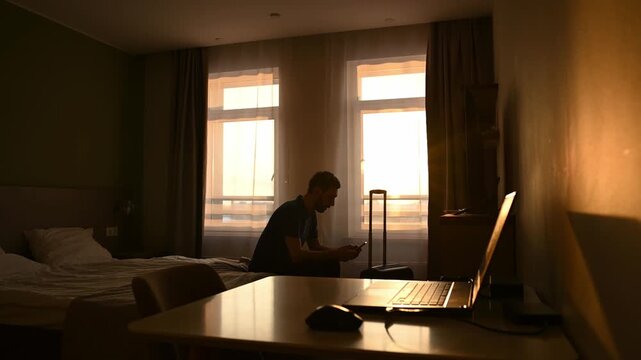 Young man finishing a phone call, standing up, taking his suitcase, and leaving his hotel room with his laptop on the desk, beautifully silhouetted against the warm light of the setting sun
