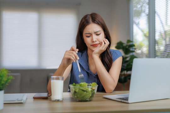 Young woman looking unhappy and bored while eating a bowl of green salad, doing a healthy diet at home