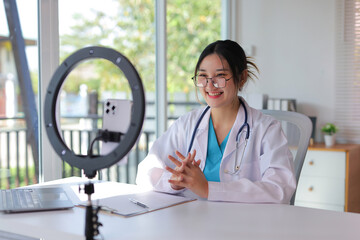 Young female doctor wearing a lab coat and stethoscope, smiling while recording a video on a...