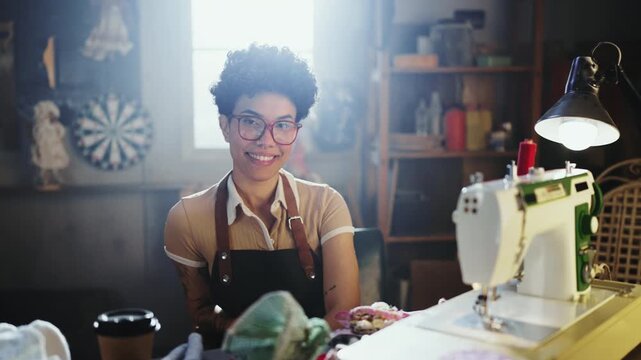 A woman works at a sewing machine in a warm, inviting workshop. She shows her skills while surrounded by fabric and tools. A coffee cup sits nearby, adding to the atmosphere.
