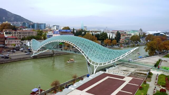 Aerial drone view of Bridge of Peace over Kura River in Tbilisi, Georgia with boats, riverfront road and urban landscape. Modern glass pedestrian bridge, cityscape, autumn trees, travel destination.