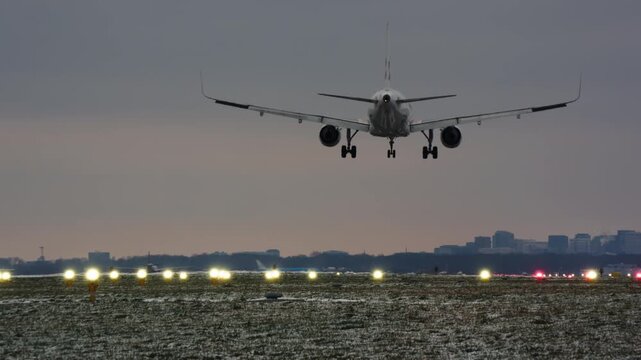 A passenger airplane lands on an illuminated runway during a cold winter evening. The shot follows the jet from final approach to touchdown zooming in to the snowy airfield with a city skyline in the
