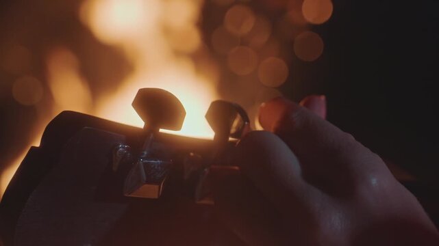 Adjusting pegs in warm light. Rustic scene shows musician preparing under fiery backdrop with hands. Camper musician sets guitar tuning in firelit ambiance before evening stage performance