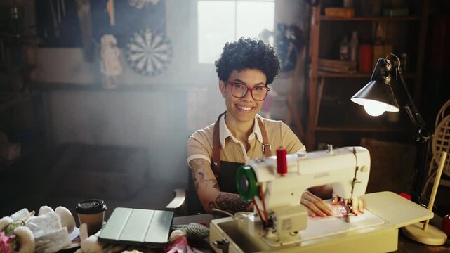 A person sits at a sewing machine in a workshop. They work on a project surrounded by fabric pieces and tools. Sunlight comes through a nearby window, creating a bright space.