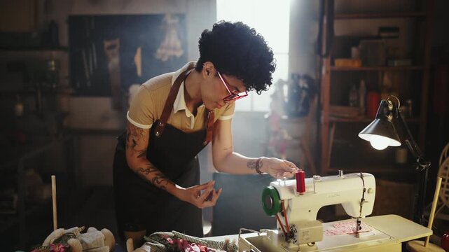 Person operates a sewing machine in a workshop. The workspace is organized with tools and materials around. The individual focuses on sewing a piece of fabric.