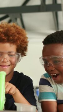 Two boys sitting table, one boy pressing cup creating green foam for science, other boy laughing
