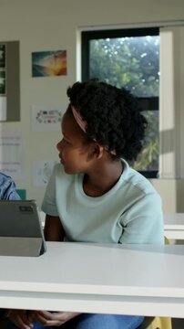 Vertical video: Noting peer, girl in mint top pink band turning left at classroom desk with tablet