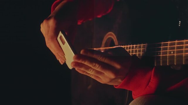 Closeup of woman tuning guitar with device. Detailed image of woman tuning guitar with phone in firelight. Woman finetunes guitar strings with phone under campfire glow while focusing closely