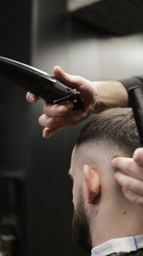 Close-up of professional barber trimming a client's hair with electric clipper. Back view of male grooming process with stylish patterned cape and neck strip in barbershop.