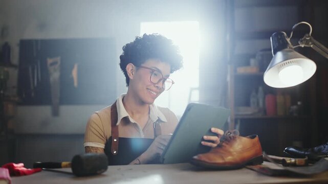 A worker in a shoe repair shop looks at a tablet. Tools and shoes are on the table. The setting shows a bright space with items around. The worker smiles while checking information.