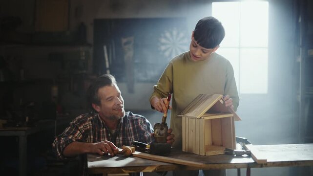 A father and son work together to build a birdhouse in a workshop. The son uses a tool to shape the roof while the father teaches him how to build it. They share smiles and learn together.