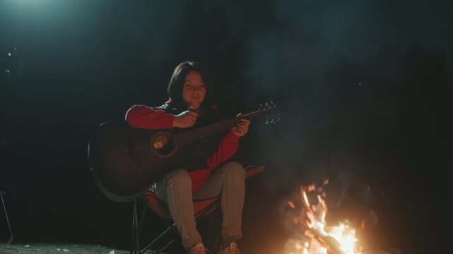 Cheerful artist strumming by glowing embers. Energetic young performer with acoustic music around pine trees. Young musician enjoying cheerful melodies beside campfire with sparks and warm glow