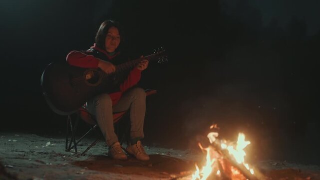 Camping guitarist warming hands by fire prepares chord progression, practices riffs, embers crackling, sandy ground, quiet night, reflective pose, distant stars, steady tempo, learning focus