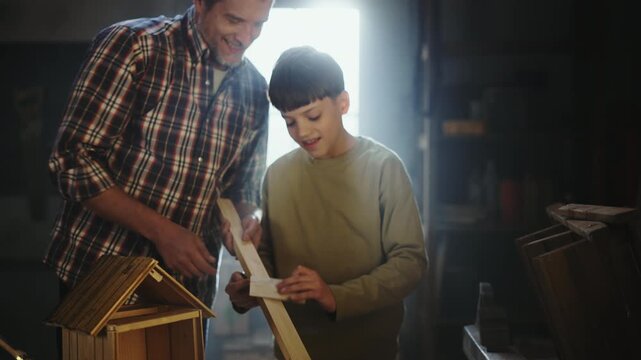 A father and son are in a workshop. They focus on a project with wooden materials. The room has light coming from a window. They share a moment of learning and teamwork.