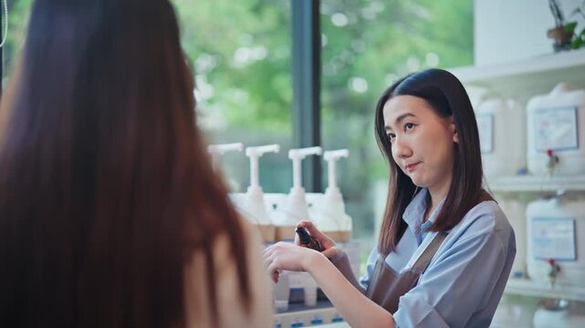 Young Asian woman in an apron holds up a small dark bottle and demonstrates a natural organic product to a customer inside a zero waste refill store.
