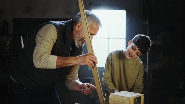Two people work together in a workshop. They cut and assemble wood pieces into a project. One is an adult and the other is a child. They focus on their work with tools and materials.