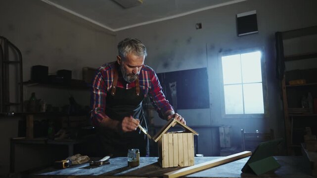 Craftsman works on a wooden birdhouse in a well-lit workshop. He measures, cuts, and paints wood to create a home for birds. Tools and materials are arranged around him.