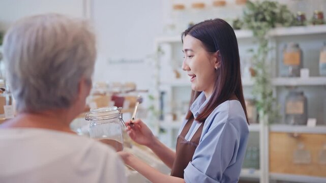 Young Asian female employee in an apron gently shows a glass jar to a senior woman while assisting her inside a warm, natural zero waste refill store.