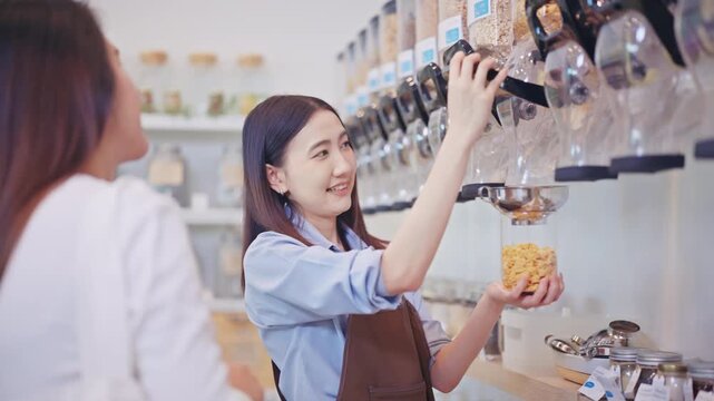 Cheerful young Asian woman in an apron operates a bulk food dispenser to serve a customer, eco-friendly refill shop stocked with natural organic products.