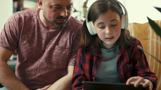 Preteen girl in headphones showing interesting content on digital tablet to her father at home-4k,10 bit footage.