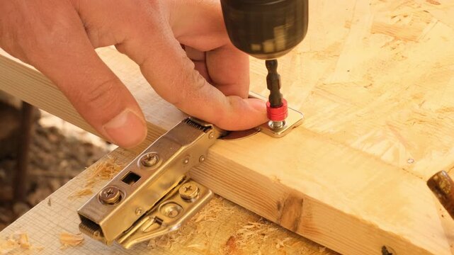 Furniture maker using electric screwdriver to attach door hinge to new cabinet. Using electric drill to screw into wooden cabinet door. Furniture production from wood. Work in woodworking shop.