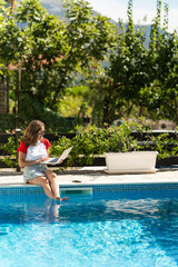 Naklejka premium Freelance woman working remotely with a laptop near a swimming pool