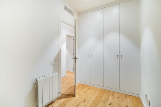 A modern, minimalistic interior of a room with white walls and wooden flooring. A closed door leads to another space, and a radiator is visible on the wall.