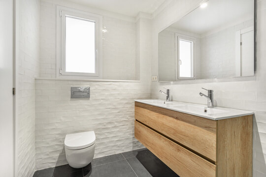 Modern bathroom with white walls, wooden vanity, and large mirror. Features a toilet and natural light from a window.