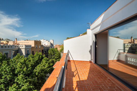 A modern balcony view in Barcelona. The scene includes a clear blue sky, terracotta tiles, and green trees lining the street. Urban architecture is visible in the background.