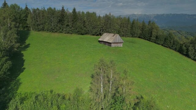 Low altitude drone flyover approaching a weathered traditional wooden barn on a hayfield, revealing the scenic Simon village valley and distant Piatra Craiului mountains, Transylvania, Romania, aerial