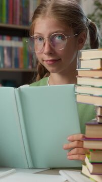 Motivated cute Caucasian little girl child school kid in glasses reading book looking at camera daughter elementary primary learning studying in library pupil reader read study learn holding textbook