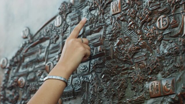 Closeup hand pointing bronze map plaque, tourist exploring textured relief with bracelet and ring, tracing route numbers and landmarks, outdoor exhibit mood with warm patina and detailed topography