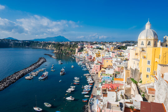 High angle view of the colorful houses and boats in the harbor of Marina Corricella on Procida Island, Italy, with the Santuario di Santa Maria delle Grazie dome.
