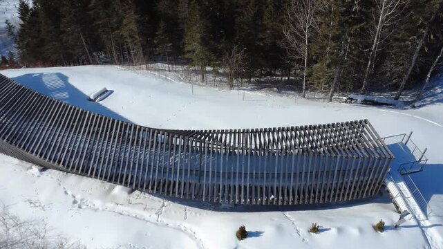 Aerial winter landscape of a curved wooden structure on a snowy hill. Overlooking the frozen Matapedia River, highway bridge, and dense forest in the Gaspe Peninsula, Quebec, Canada.
