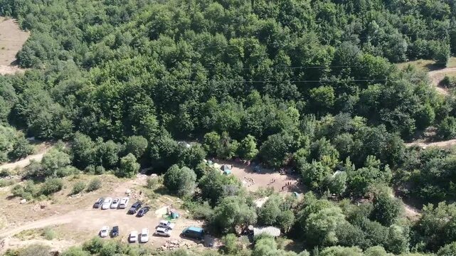 Aerial view of a lively summer camp in a forest clearing. People are playing a game among colorful tents surrounded by lush green trees on a sunny day.