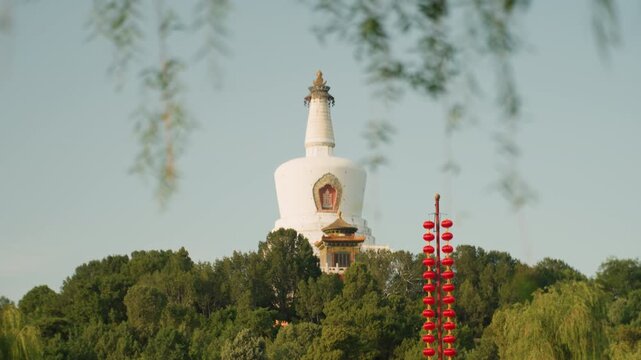 White stupa framed by willow branches rising above dense green trees with red ceremonial tower in foreground clear blue sky and soft sunlight create tranquil mindful travel mood, showcasing buddhist
