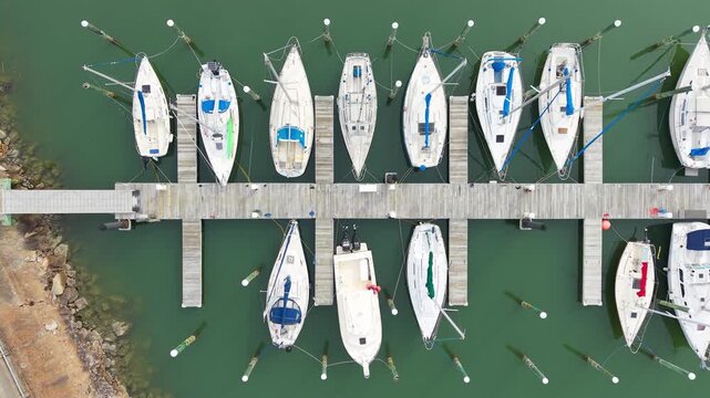 Aerial top-down view of various sailboats and motorboats docked in wooden slips at a calm coastal marina during the day.