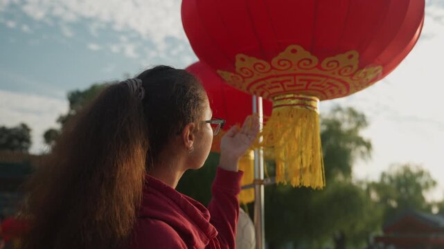 Black girl inspects red lantern outdoors in golden hour park, backlit profile reaching to touch silk tassel, wearing hoodie and glasses, long curly ponytail, reflective mood, cultural festival