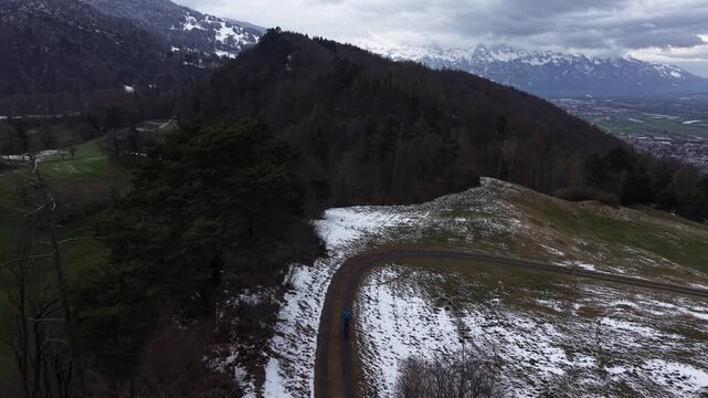 Drone following cyclist riding downhill on winding mountain road with patches of snow and valley views. Outdoor adventure and mobility in alpine spring conditions