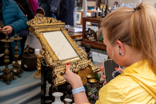 A woman examines an ornate vintage mirror among assorted antiques and porcelain on a crowded outdoor flea market table