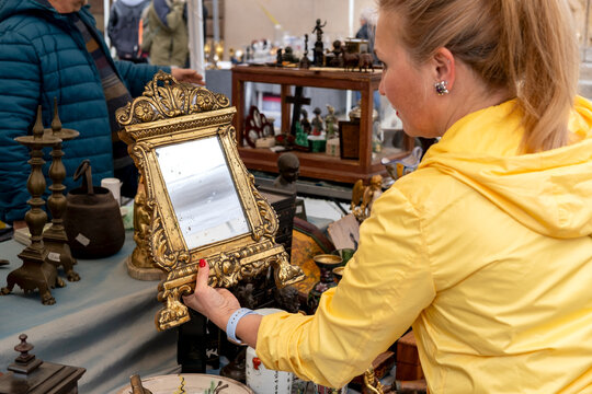 A woman examines an ornate vintage mirror among assorted antiques and porcelain on a crowded outdoor flea market table