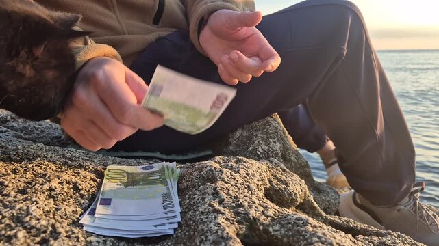 Money counting euros, man counts stack of 100 euro banknotes sitting by the sea at sunrise, symbolizing savings and financial planning